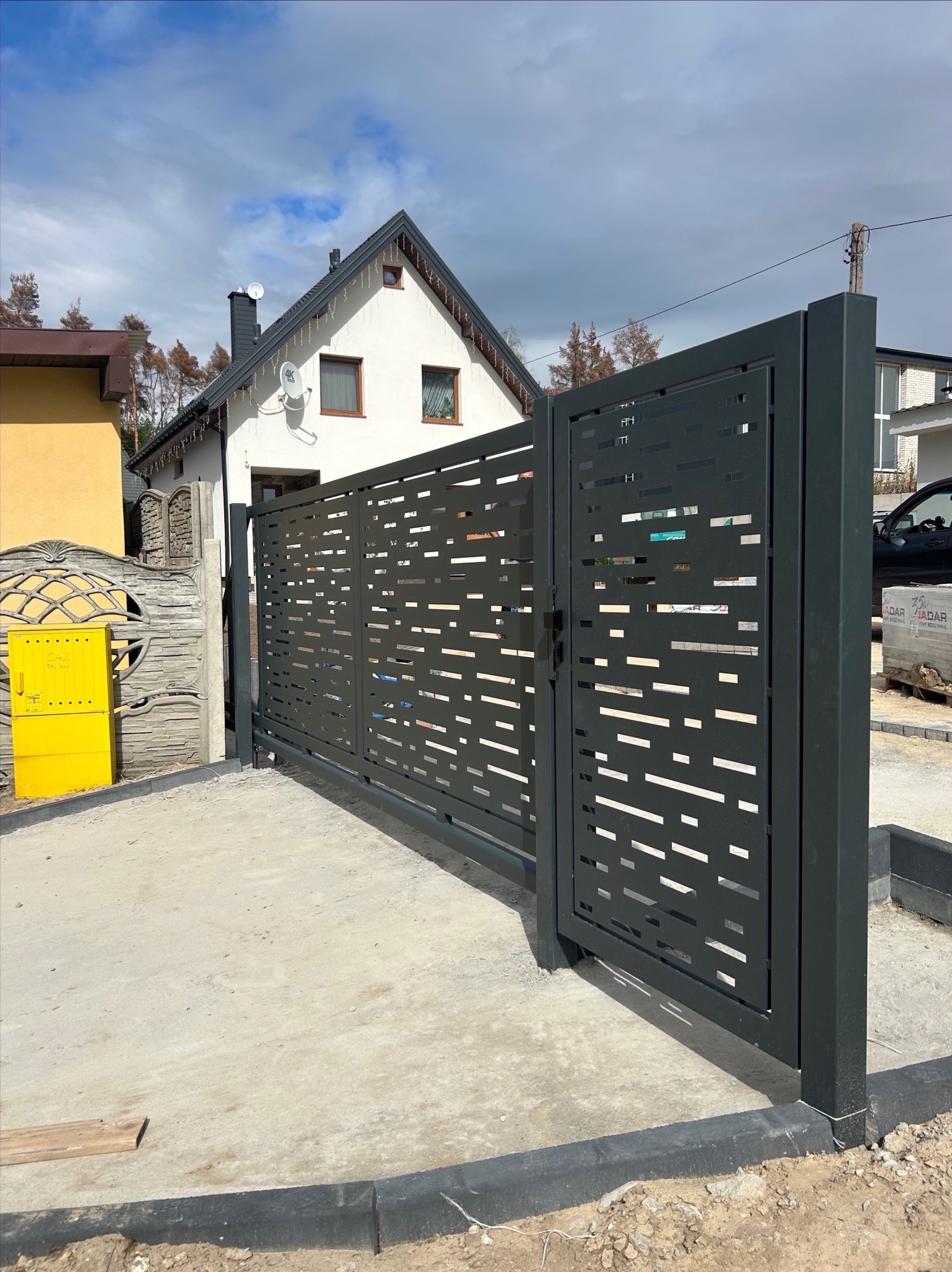 Nowoczesna, przesuwna brama wjazdowa z ażurowym wzorem, pomalowana na grafitowo. Widoczny fragment domu z białą elewacją i żółta skrzynka gazowa.