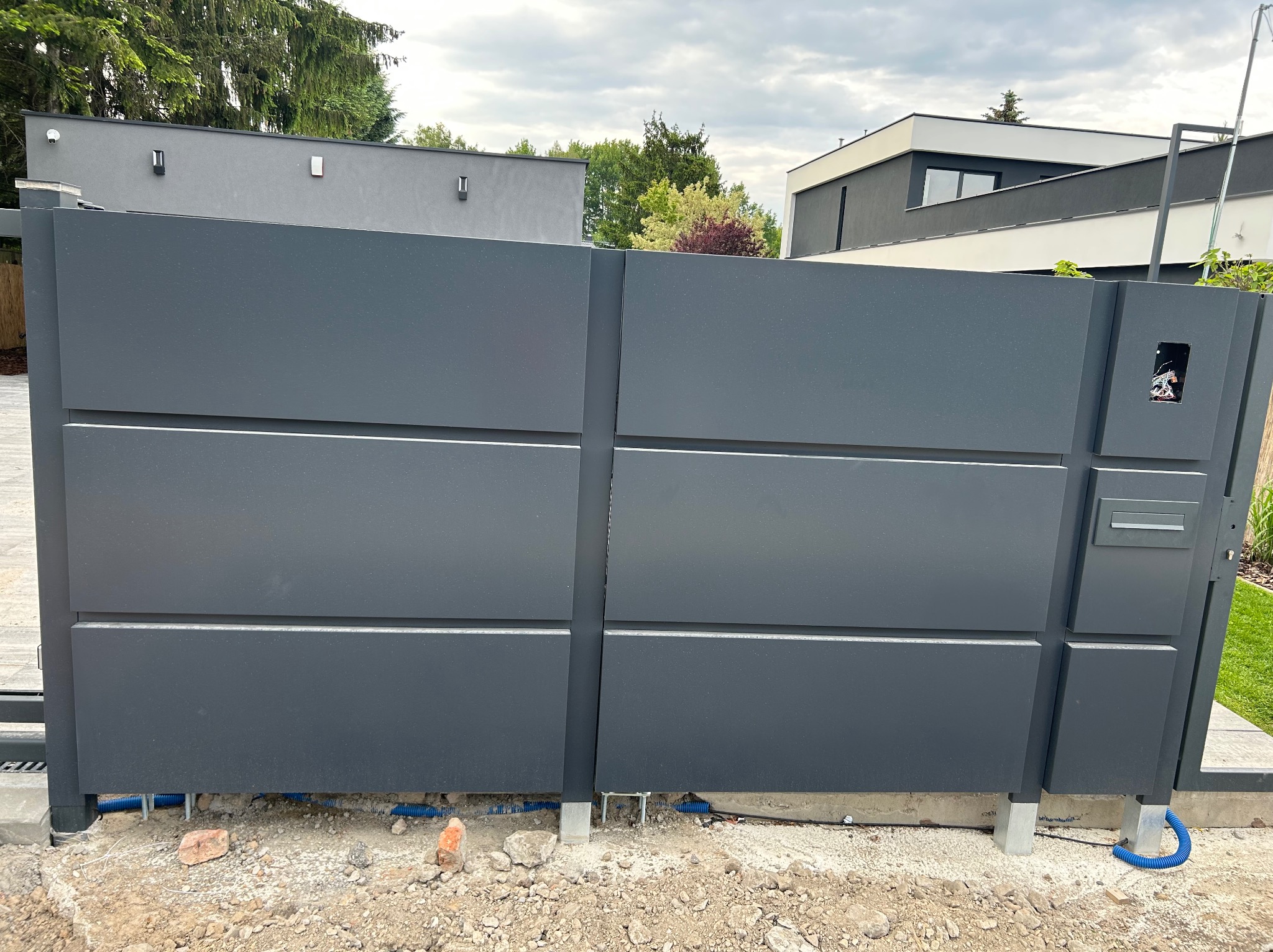 Nowoczesna, przesuwna brama wjazdowa w kolorze grafitowym z widoczną skrzynką na listy i instalacją elektryczną w ziemi. Tło stanowią budynki mieszkalne i zieleń.