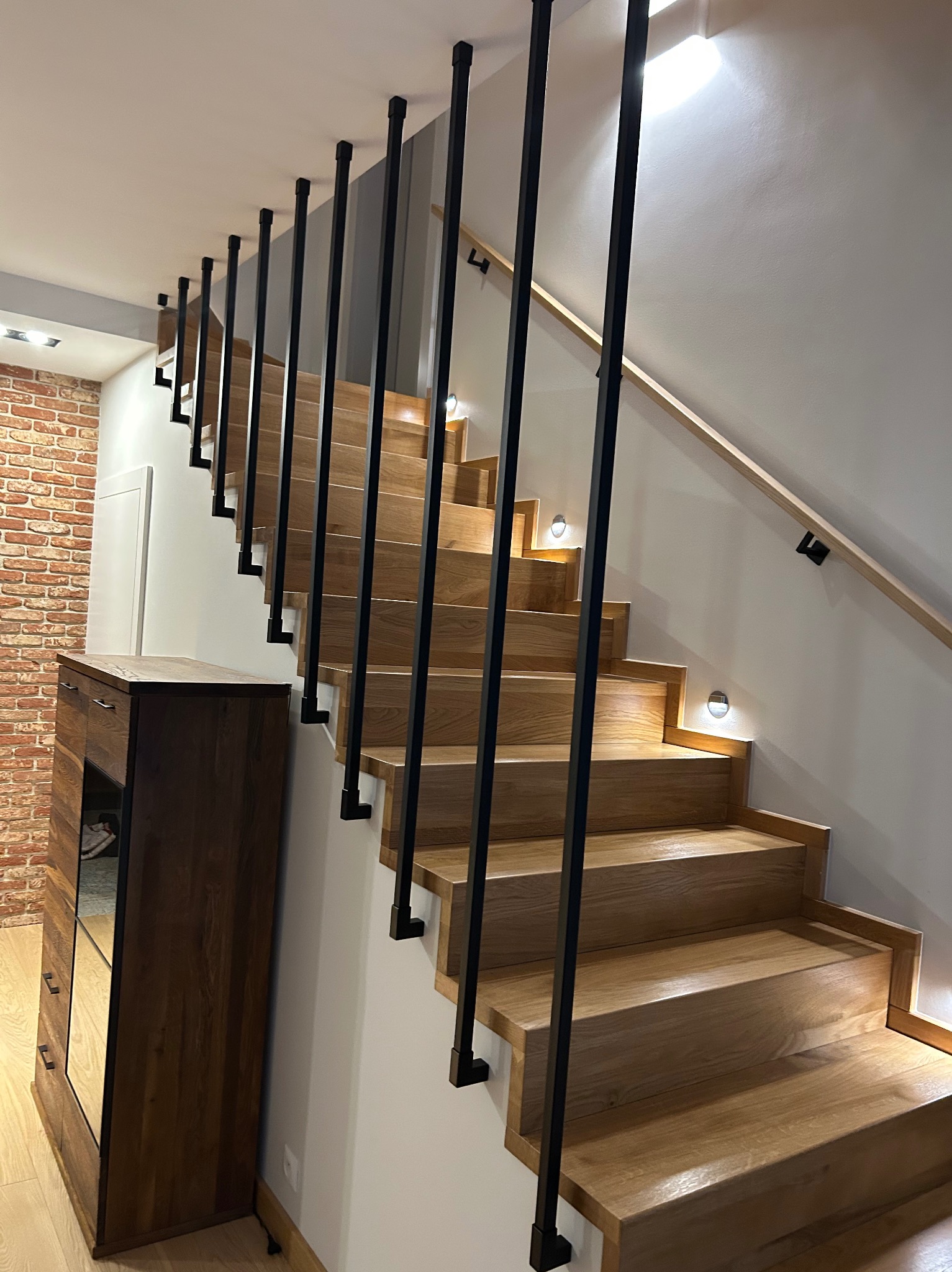 Drewniane schody z czarną, metalową balustradą w nowoczesnym wnętrzu. Stopnie oświetlone punktowymi lampami. Widoczna część ściany z cegły i drewniana komoda.