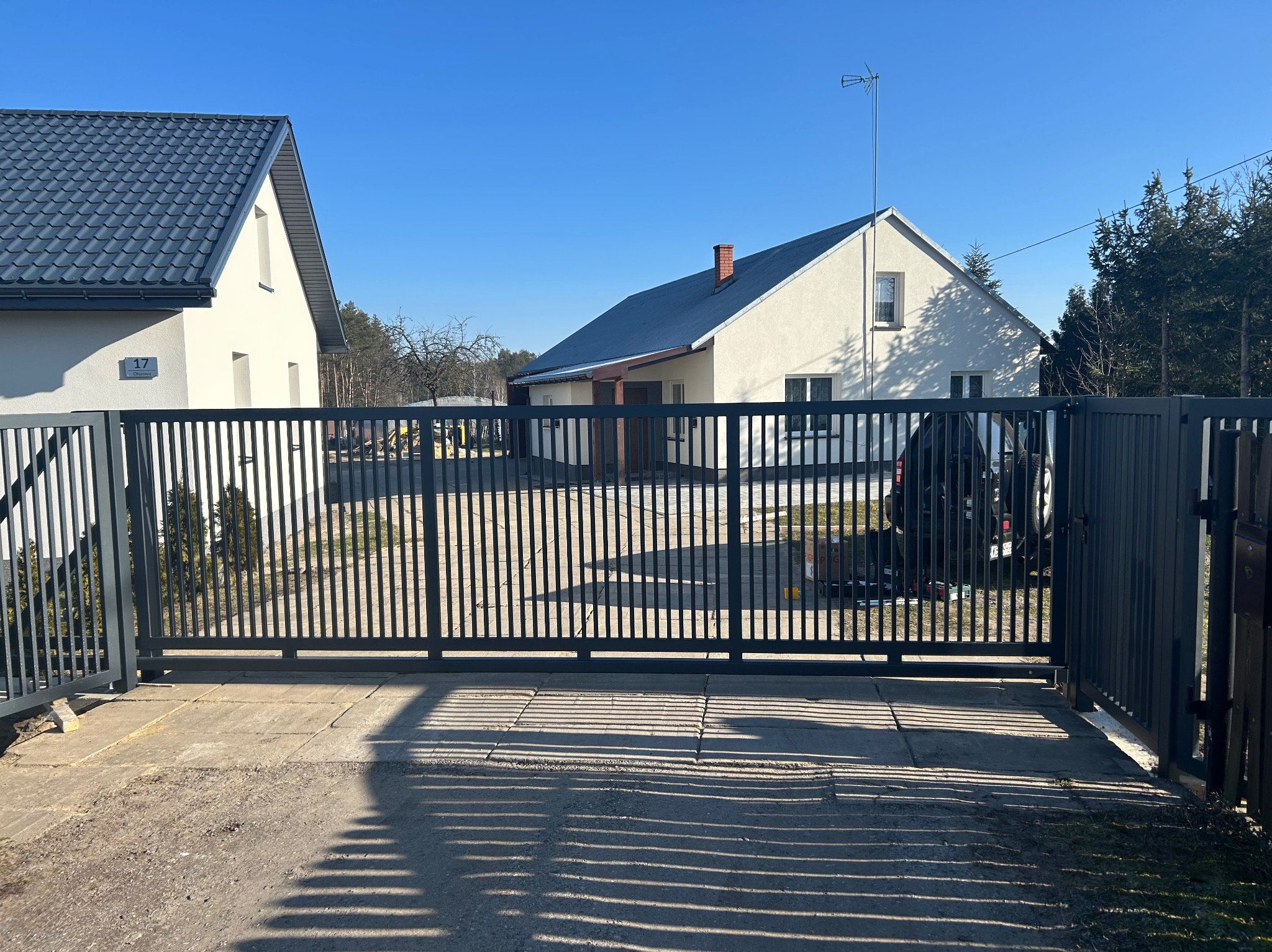 Nowoczesna, przesuwna brama wjazdowa w kolorze grafitowym, z pionowymi szczeblami, na tle jasnego domu z ciemnym dachem i błękitnego nieba w słoneczny dzień.