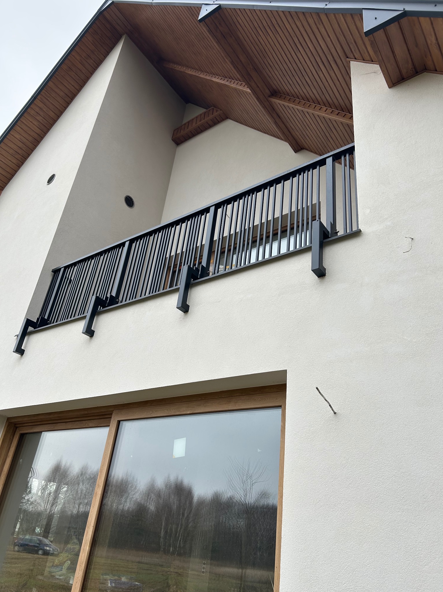 Czarna, metalowa balustrada balkonowa z pionowymi szczeblami, zamontowana na jasnej elewacji nowoczesnego domu. Widok z dołu, okno z drewnianą ramą w tle.