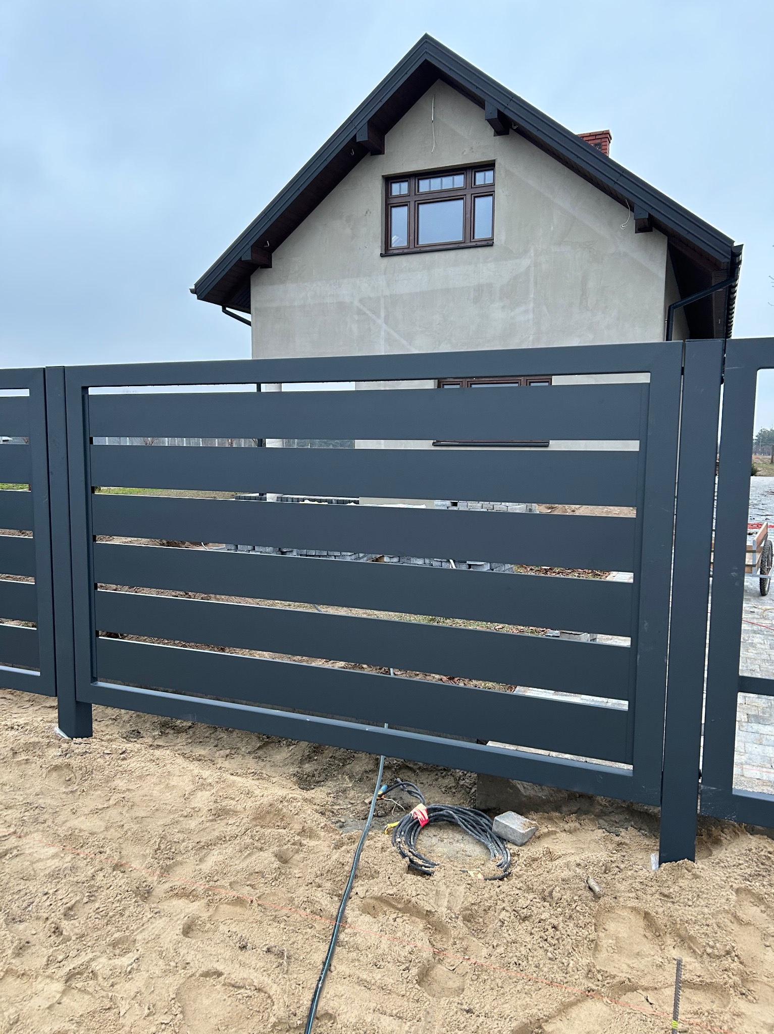 Nowoczesna, przesuwna brama wjazdowa z poziomymi panelami w kolorze grafitowym, na tle domu w trakcie budowy. Widoczne przewody elektryczne przy fundamencie.