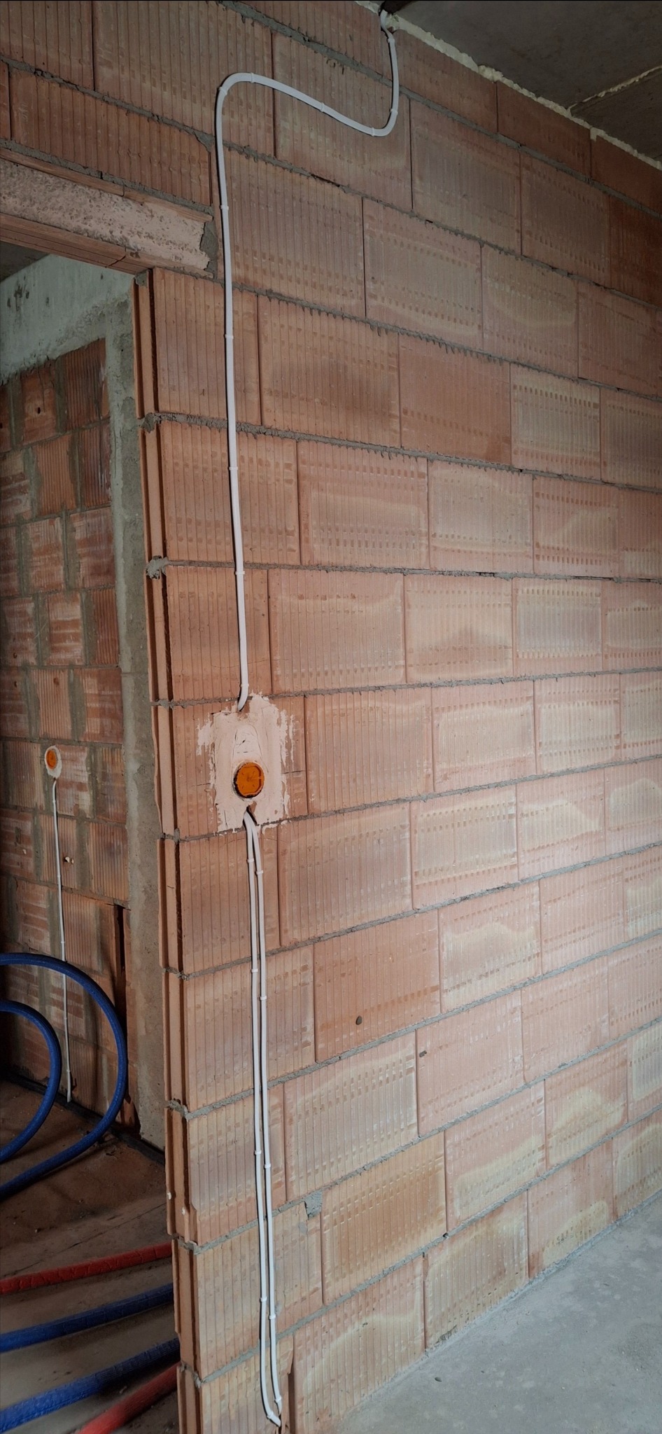 Białe kable elektryczne prowadzone po ścianie z cegły dziurawki, z widoczną puszką instalacyjną z pomarańczowym wnętrzem, w tle fragment instalacji hydraulicznej.