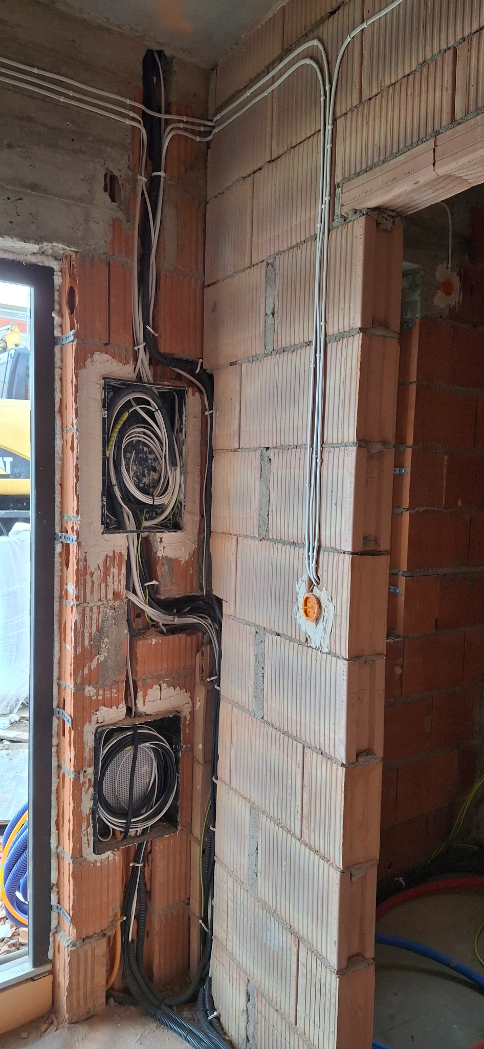 Wewnętrzna instalacja elektryczna w budynku w trakcie budowy, widoczne przewody w ścianach z cegły, puszki instalacyjne, peszle i kable prowadzone po ścianach.