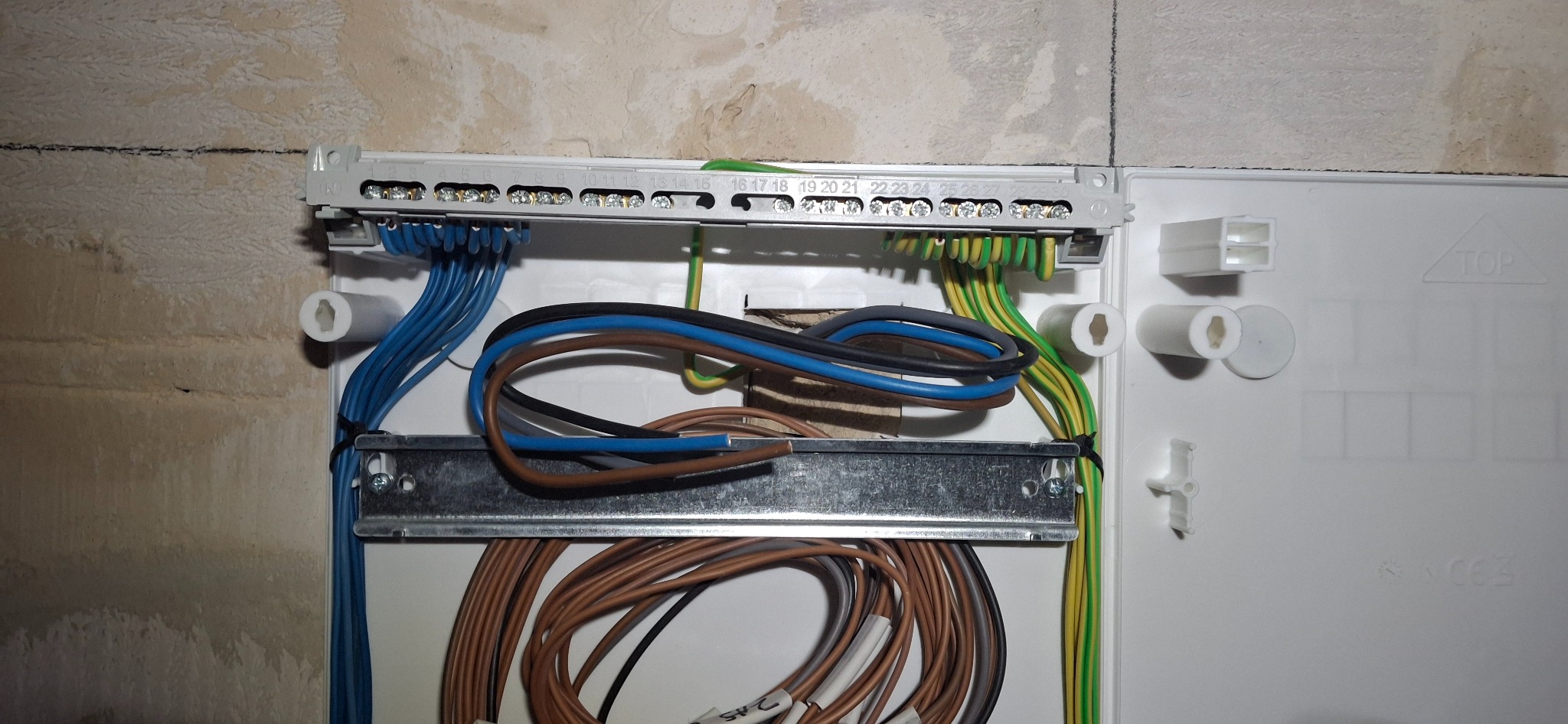 Rozdzielnica elektryczna w trakcie montażu, widoczne kable w różnych kolorach (niebieski, brązowy, zielono-żółty) podłączone do listwy zaciskowej, tło ściana w trakcie wykończenia.