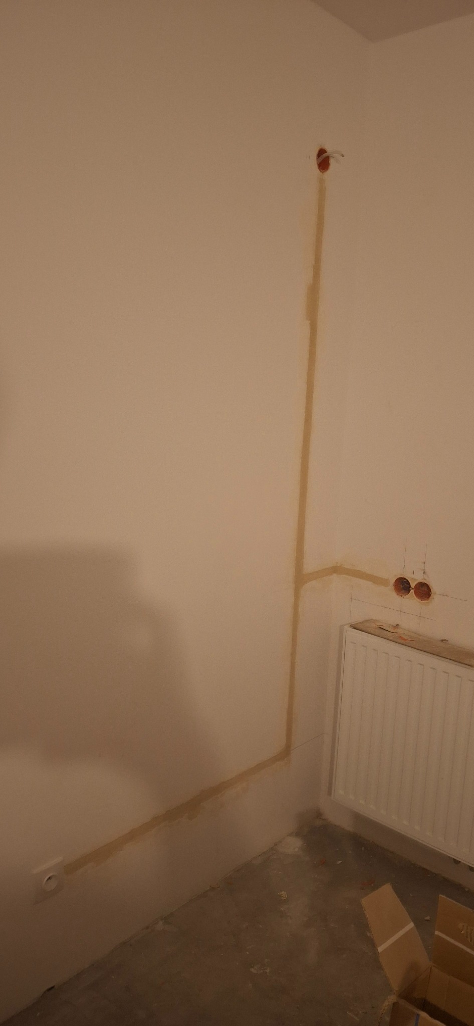 Ściana z wyfrezowanymi bruzdami pod przewody elektryczne, zabezpieczonymi gipsem, widoczne gniazdko oraz puszki instalacyjne w ścianie, obok grzejnik.