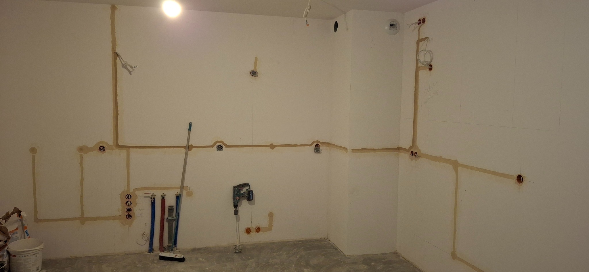Ściana w trakcie bruzdowania pod przewody elektryczne, widoczne wyżłobienia wypełnione gipsem, puszki instalacyjne, wiertarka z mieszadłem do gipsu oparta o ścianę, rury instalacyjne i worek...