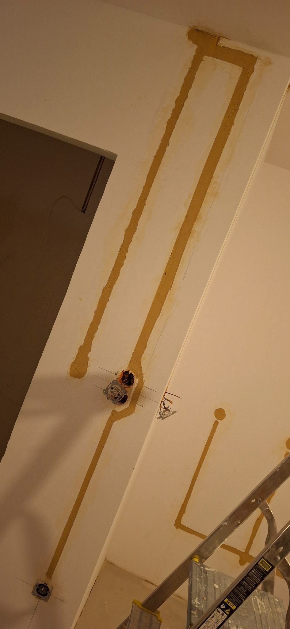 Wykute bruzdy w ścianie pod przewody elektryczne, widoczne puszki pod gniazdka i włączniki, częściowo wypełnione gipsem, drabina aluminiowa w rogu kadru.