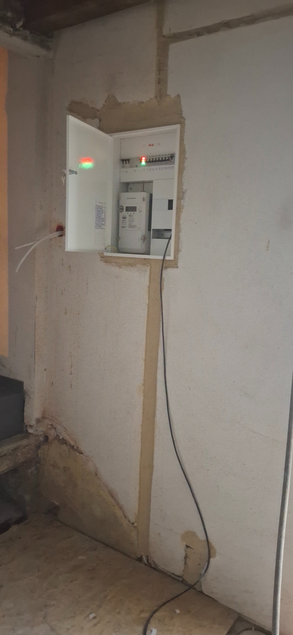 Otwarta skrzynka elektryczna z licznikiem i zabezpieczeniami w surowym wnętrzu, widoczne kable i niedokończone wykończenie ściany.