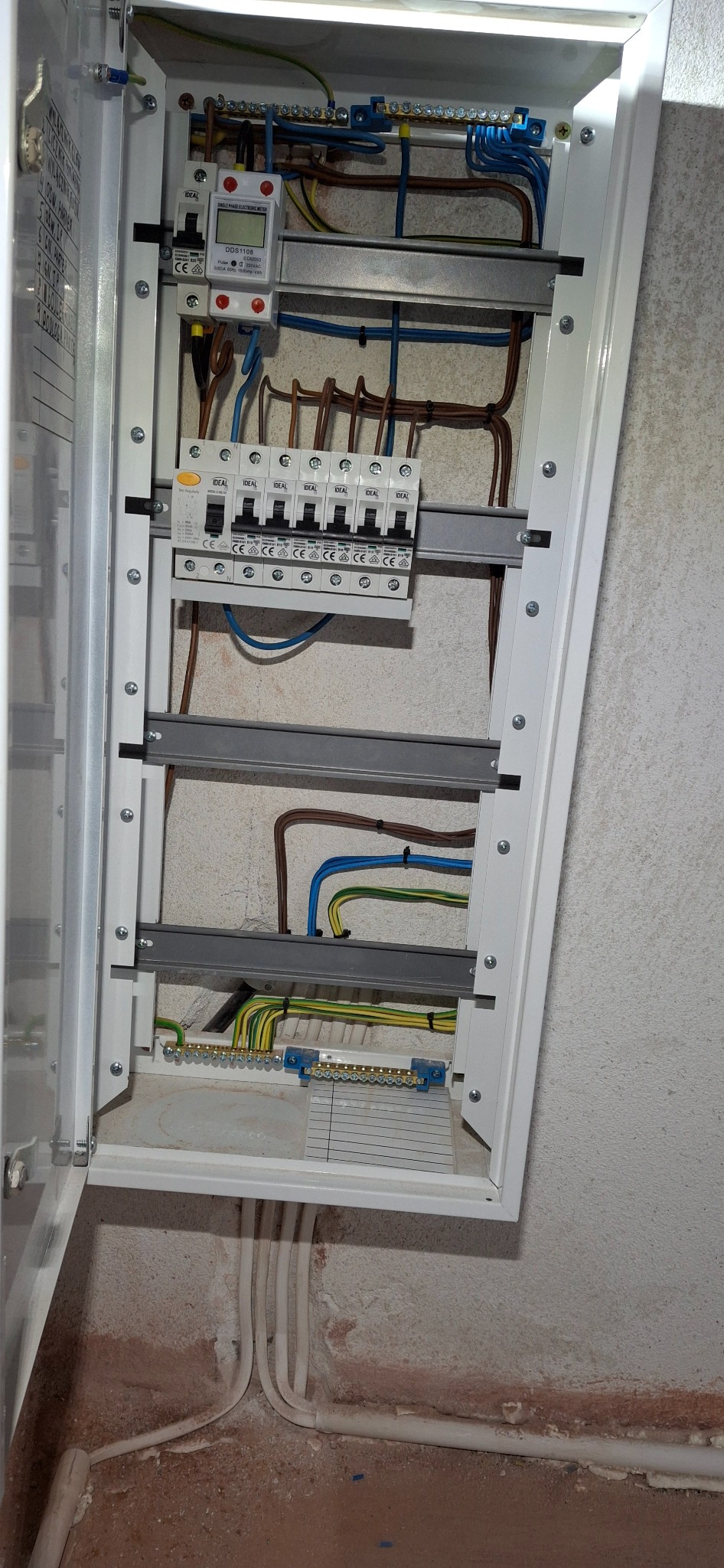 Otwarta skrzynka elektryczna z widocznymi bezpiecznikami, licznikiem energii oraz przewodami w różnych kolorach, umieszczona na ścianie z widoczną instalacją elektryczną w podłodze.