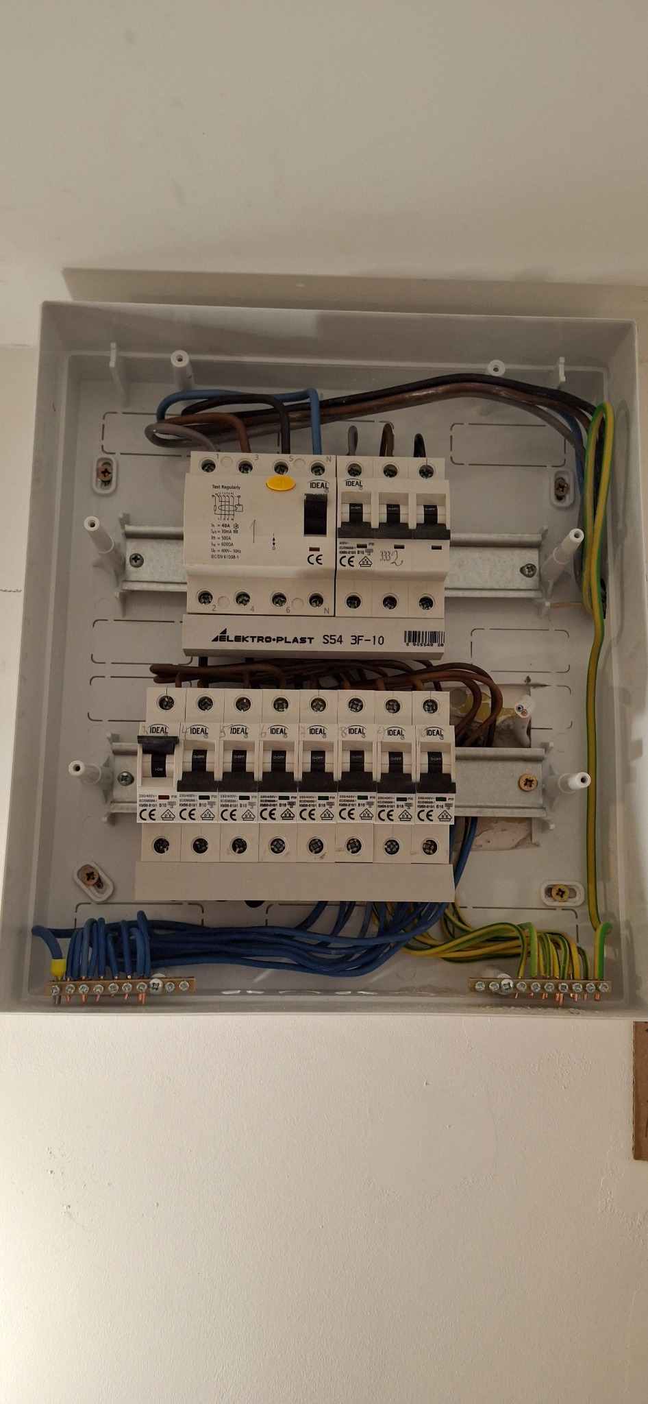 Otwarta skrzynka z bezpiecznikami firmy Elektro-Plast S54 3F-10 z widocznymi przewodami i listwami zaciskowymi, zamontowana na białej ścianie.
