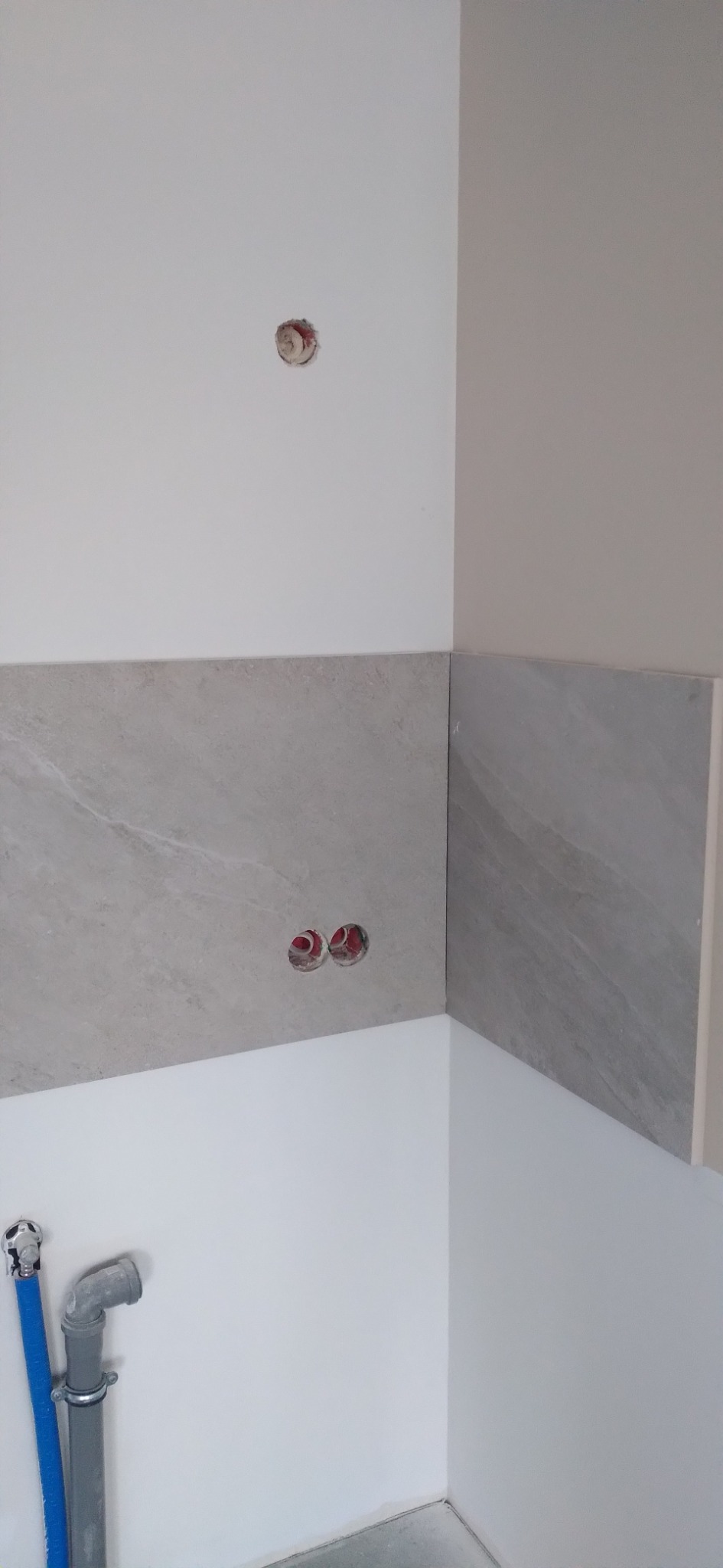 Świeżo wykafelkowana ściana z dwoma puszkami elektrycznymi bez osprzętu i otworem na górze, widoczne rury kanalizacyjne z niebieską rurką doprowadzającą wodę.