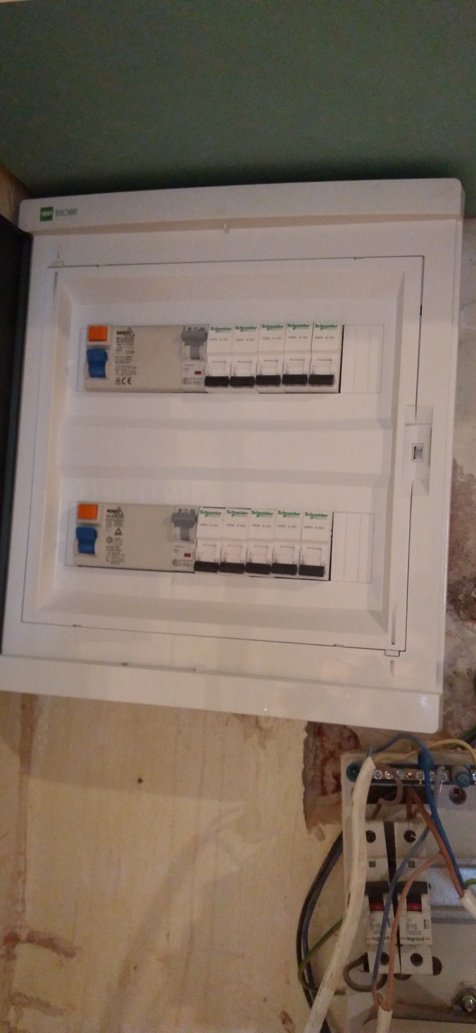 Biała skrzynka z bezpiecznikami firmy Schneider Electric zamontowana na nierównej ścianie, widoczne przewody elektryczne i zielona ściana w tle.