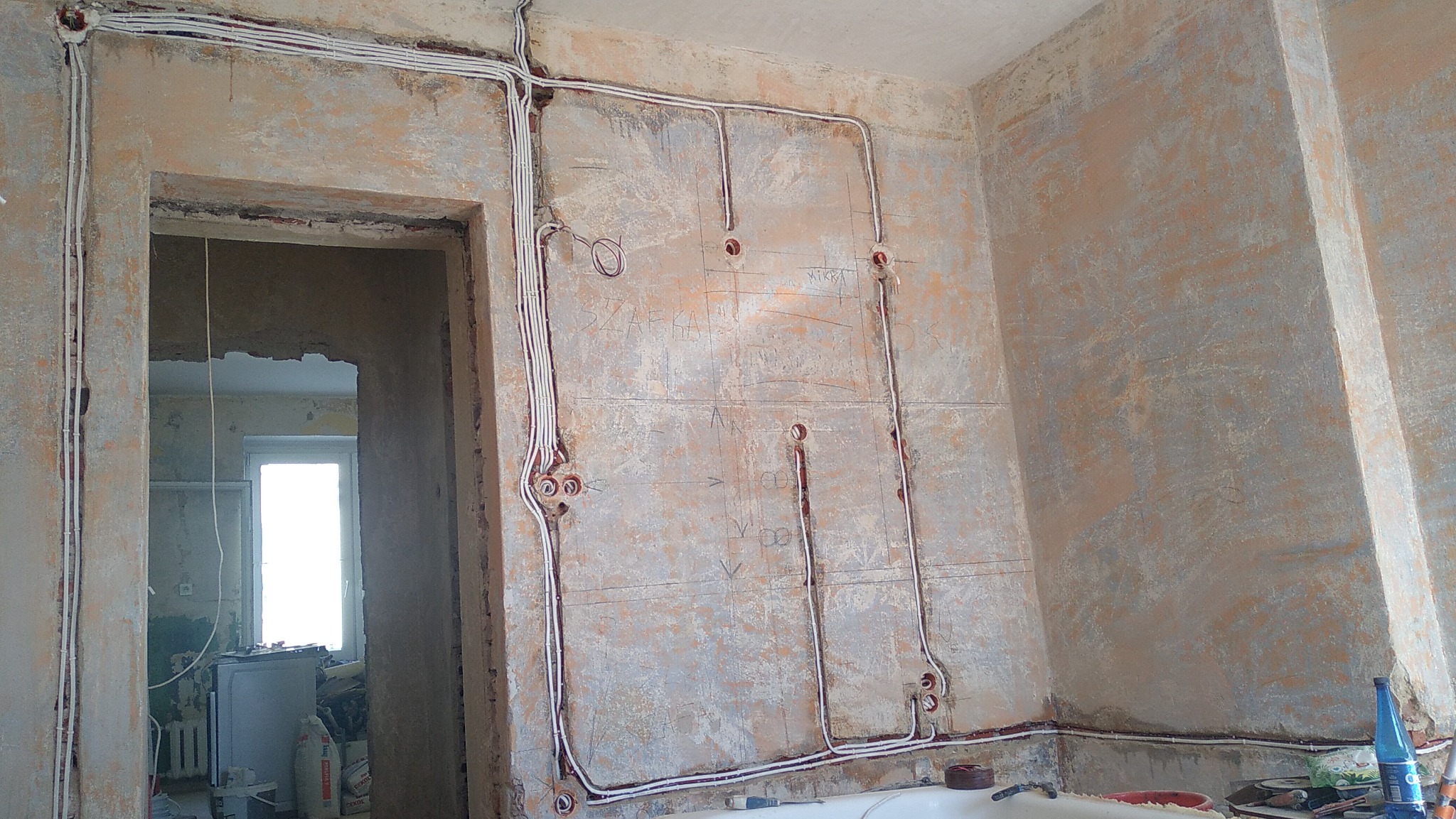 Wnętrze w trakcie remontu, widoczne poprowadzone kable elektryczne po ścianach, z zaznaczonymi punktami na gniazdka i włączniki, surowe ściany z widocznymi śladami po wcześniejszych instalacjach.