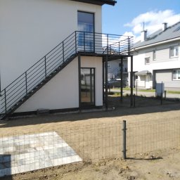 ART-SPAW - Zewnętrzne schody metalowe z balustradą prowadzące na balkon w nowoczesnym domu, widoczne fragmenty ogrodzenia panelowego i kostki brukowej na posesji.
