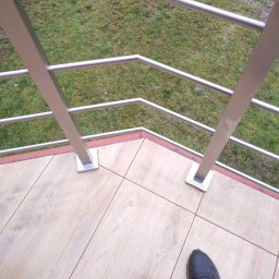 ART-SPAW - Szczegółowe ujęcie fragmentu balkonu z balustradą ze stali nierdzewnej, widok z góry na stalowe słupki i poręcze, płytki podłogowe imitujące drewno oraz fragment buta w rogu kadru.