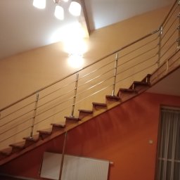 ART-SPAW - Drewniane schody z metalową balustradą na tle pomarańczowej ściany, widoczne oświetlenie sufitowe i okno z zasłoną.