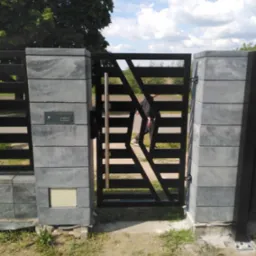 Nowoczesna brama wjazdowa z poziomymi przęsłami i geometrycznym wzorem, osadzona na szarych, betonowych słupkach z zamontowaną skrzynką na listy, widziana z perspektywy podwórza.