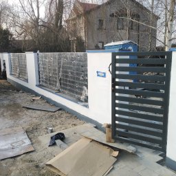 ART-SPAW - Nowoczesne ogrodzenie z bramą w trakcie budowy: białe słupki z szarymi panelami wypełnionymi folią ochronną, szara brama z poziomych profili, widoczne materiały budowlane i teren po budowie.