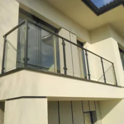 Balkon z nowoczesną balustradą szklaną z czarnymi elementami mocującymi, widoczny z dołu na tle jasnej elewacji budynku.