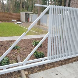 ART-SPAW - Szara, metalowa brama przesuwna w trakcie montażu, widoczne elementy konstrukcyjne i podkładki stabilizujące, tło z zielenią i elementami architektury ogrodowej.