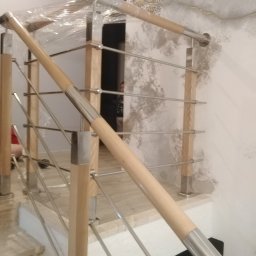 ART-SPAW - Fragment drewniano-metalowej balustrady schodowej z elementami zabezpieczonymi folią ochronną, widoczne jasne drewniane słupki i stalowe wypełnienie, tło stanowi wzorzysta ściana.