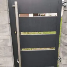 Nowoczesna, antracytowa furtka z poziomymi szczelinami i długim, pionowym uchwytem ze stali nierdzewnej, zamontowana między betonowymi słupkami ogrodzeniowymi.