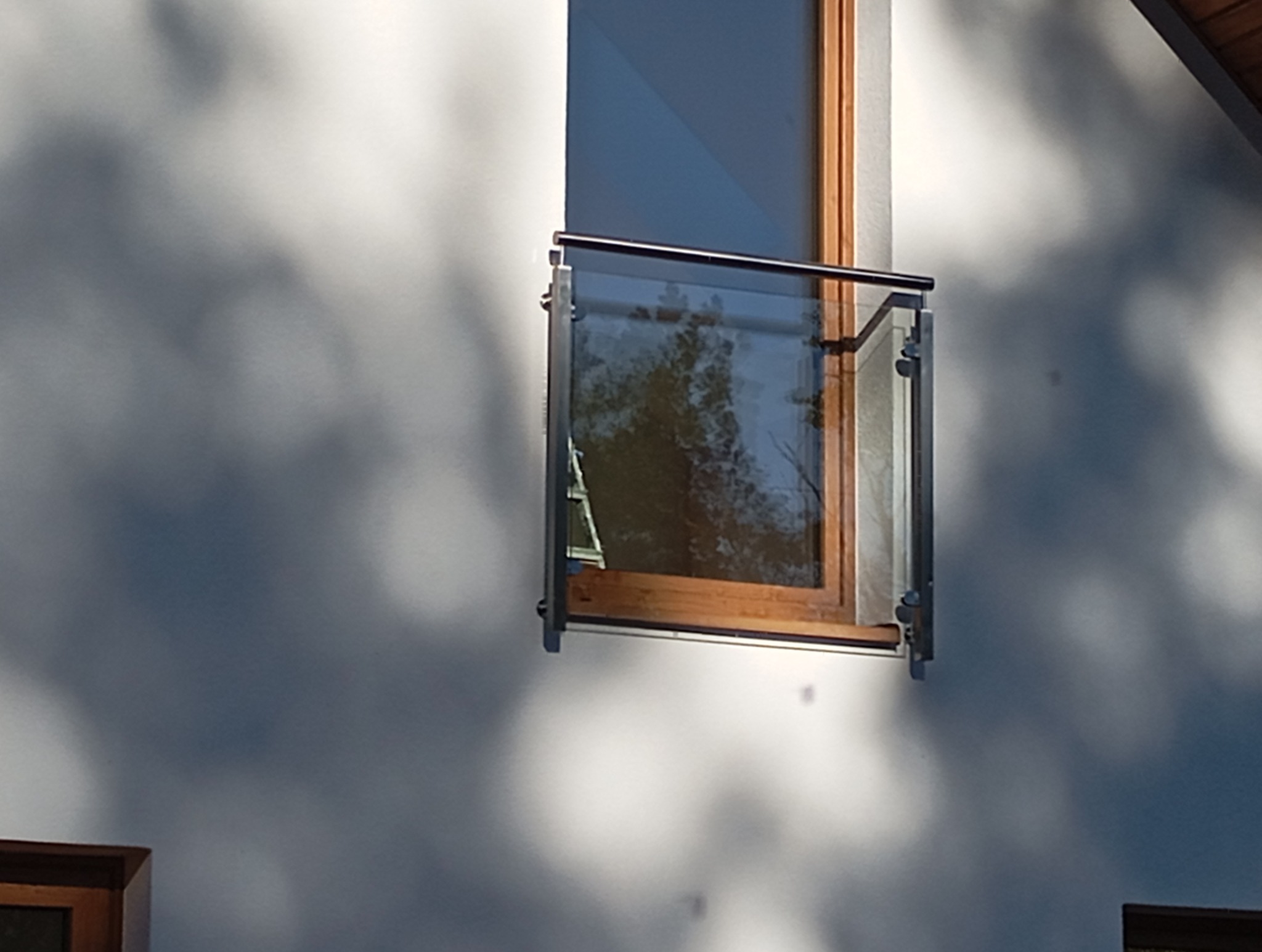 Szklana balustrada balkonowa z nierdzewnymi elementami mocującymi, zamontowana przy oknie, z widocznym odbiciem drzew w szybie i gra światła na fasadzie budynku.