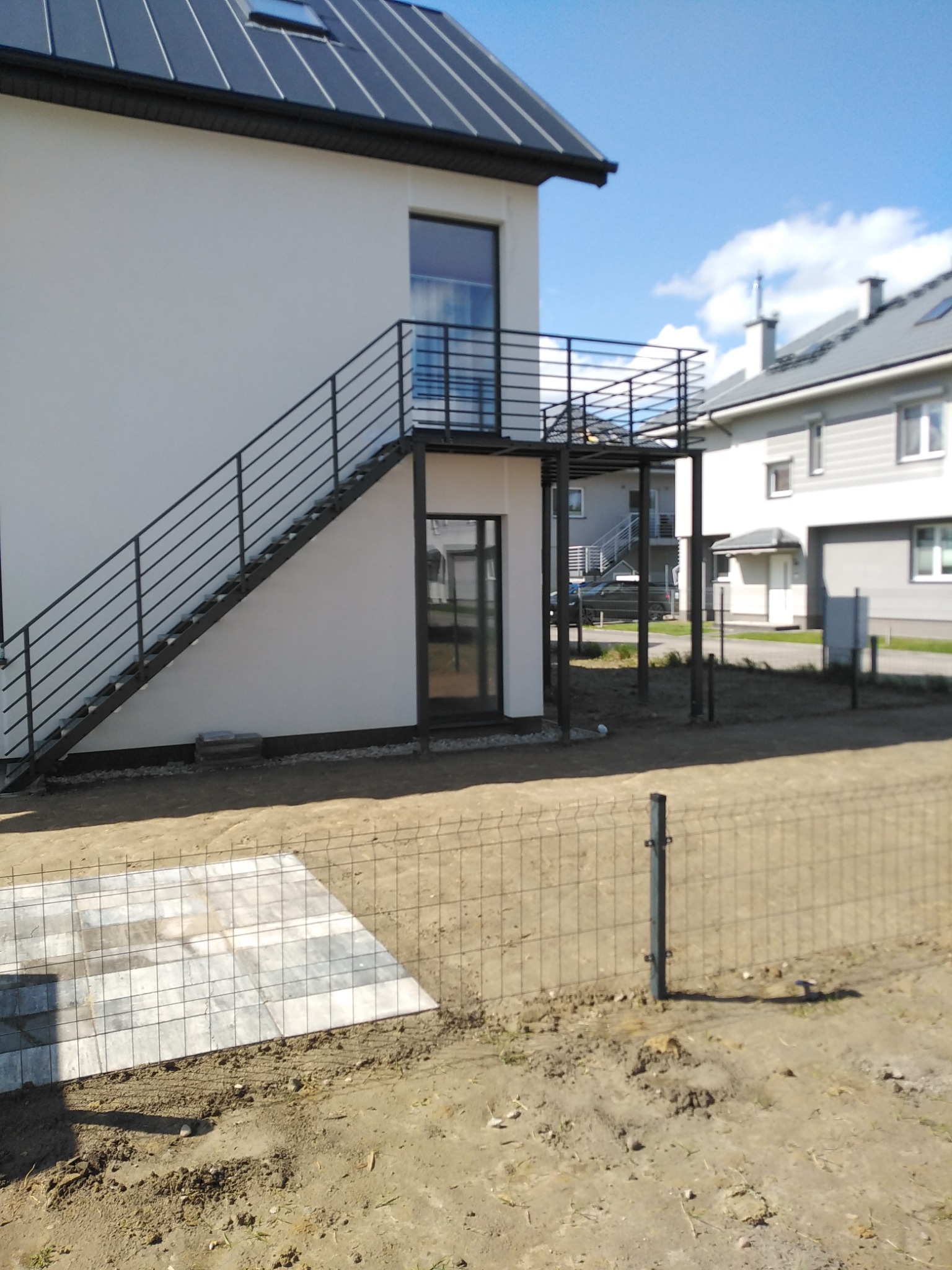 Zewnętrzne schody metalowe z balustradą prowadzące na balkon w nowoczesnym domu, widoczne fragmenty ogrodzenia panelowego i kostki brukowej na posesji.