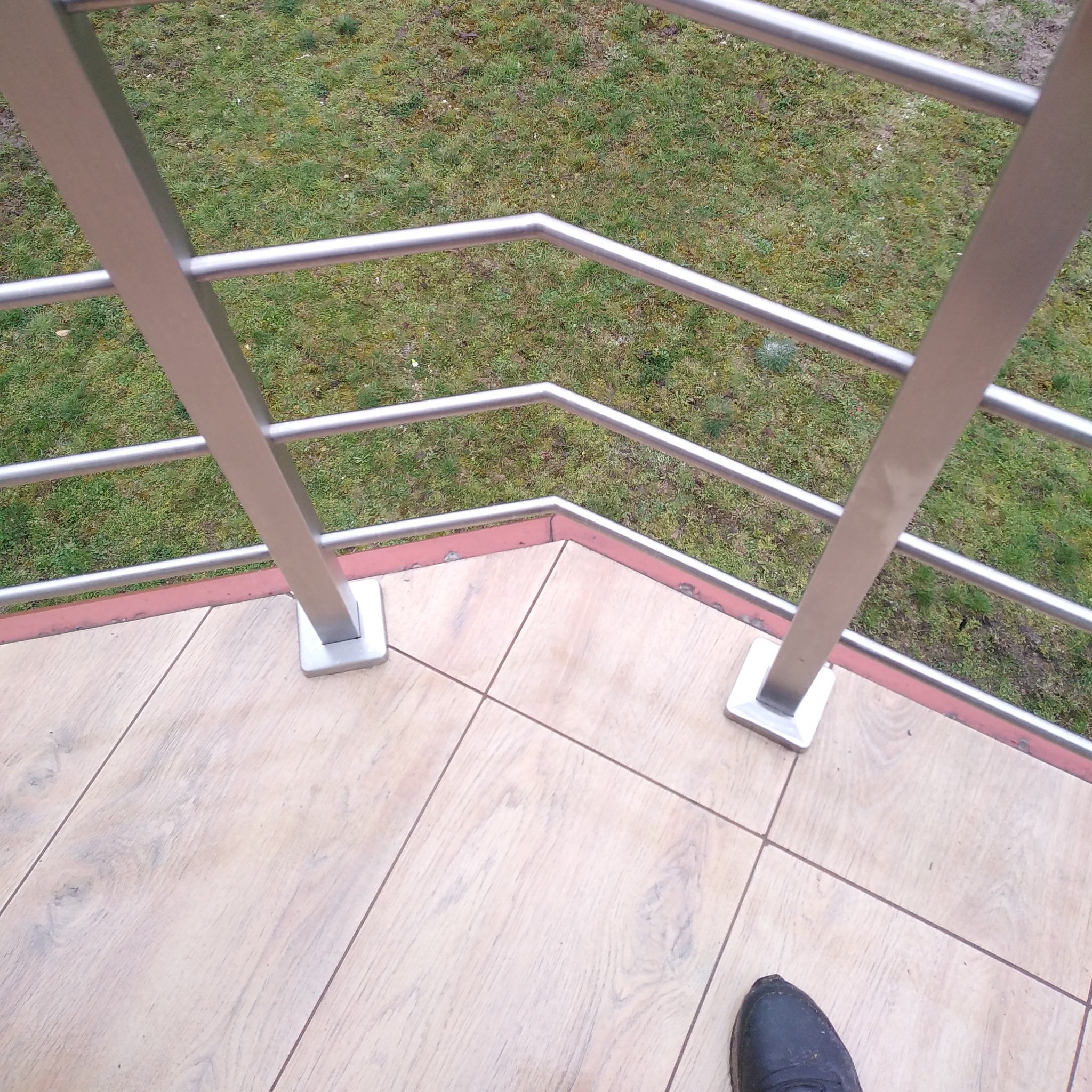 Szczegółowe ujęcie fragmentu balkonu z balustradą ze stali nierdzewnej, widok z góry na stalowe słupki i poręcze, płytki podłogowe imitujące drewno oraz fragment buta w rogu kadru.