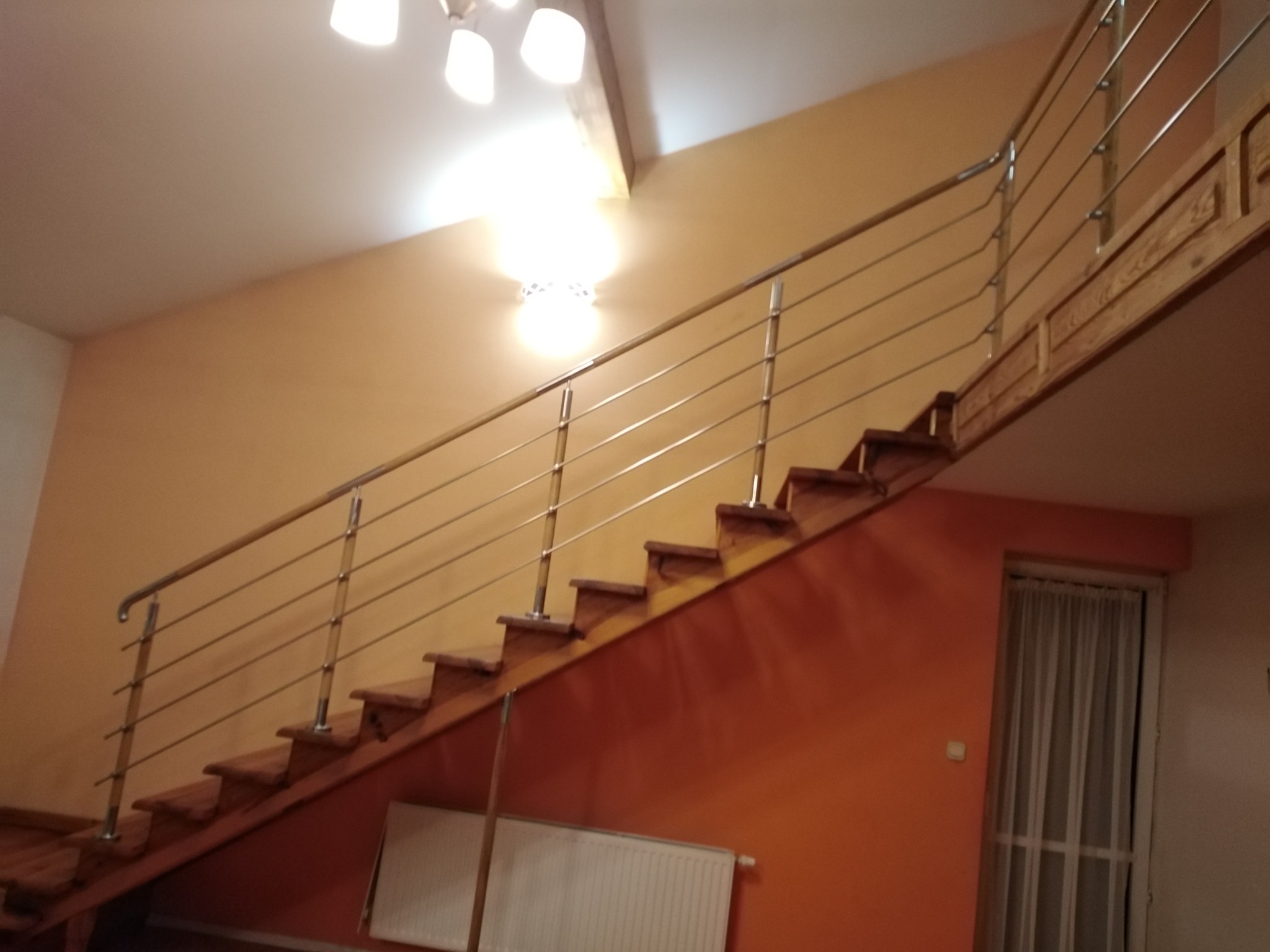 Drewniane schody z metalową balustradą na tle pomarańczowej ściany, widoczne oświetlenie sufitowe i okno z zasłoną.