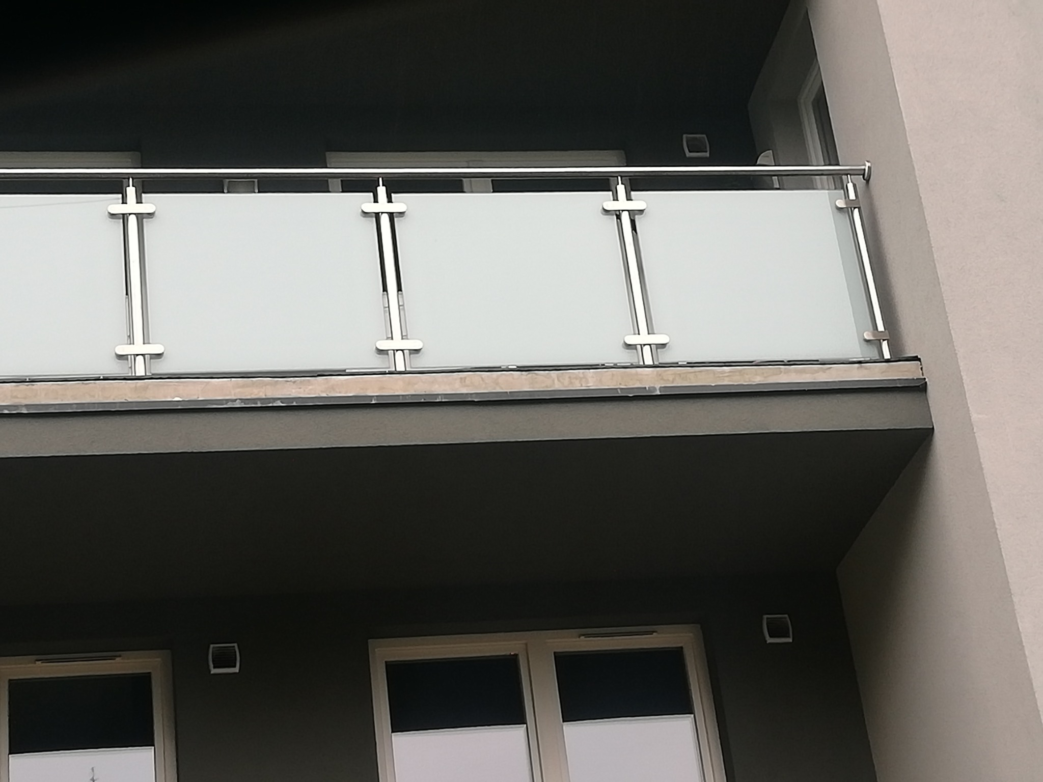 Nowoczesna balustrada balkonowa z matowego szkła i stali nierdzewnej, zamontowana na tle szarej elewacji budynku wielorodzinnego.