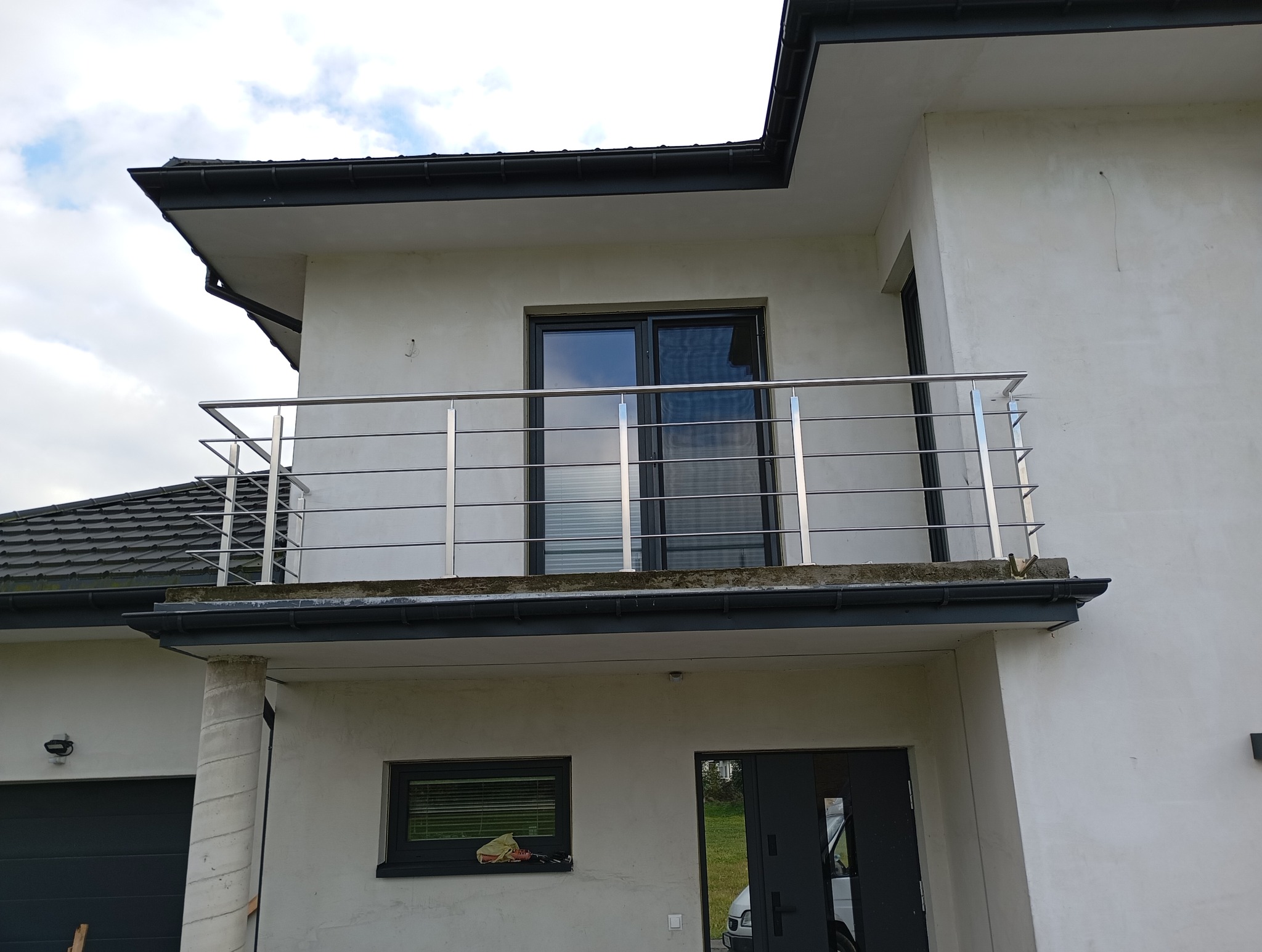 Balkon z balustradą nierdzewną w nowoczesnym domu z szarą stolarką okienną i drzwiami, widok z dołu na tle pochmurnego nieba.