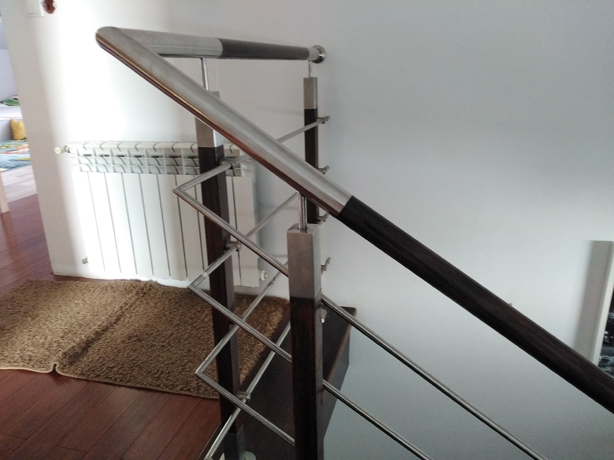 Nowoczesna balustrada ze stali nierdzewnej z drewnianymi elementami na schodach wewnątrz domu, widoczny fragment grzejnika i dywanu.