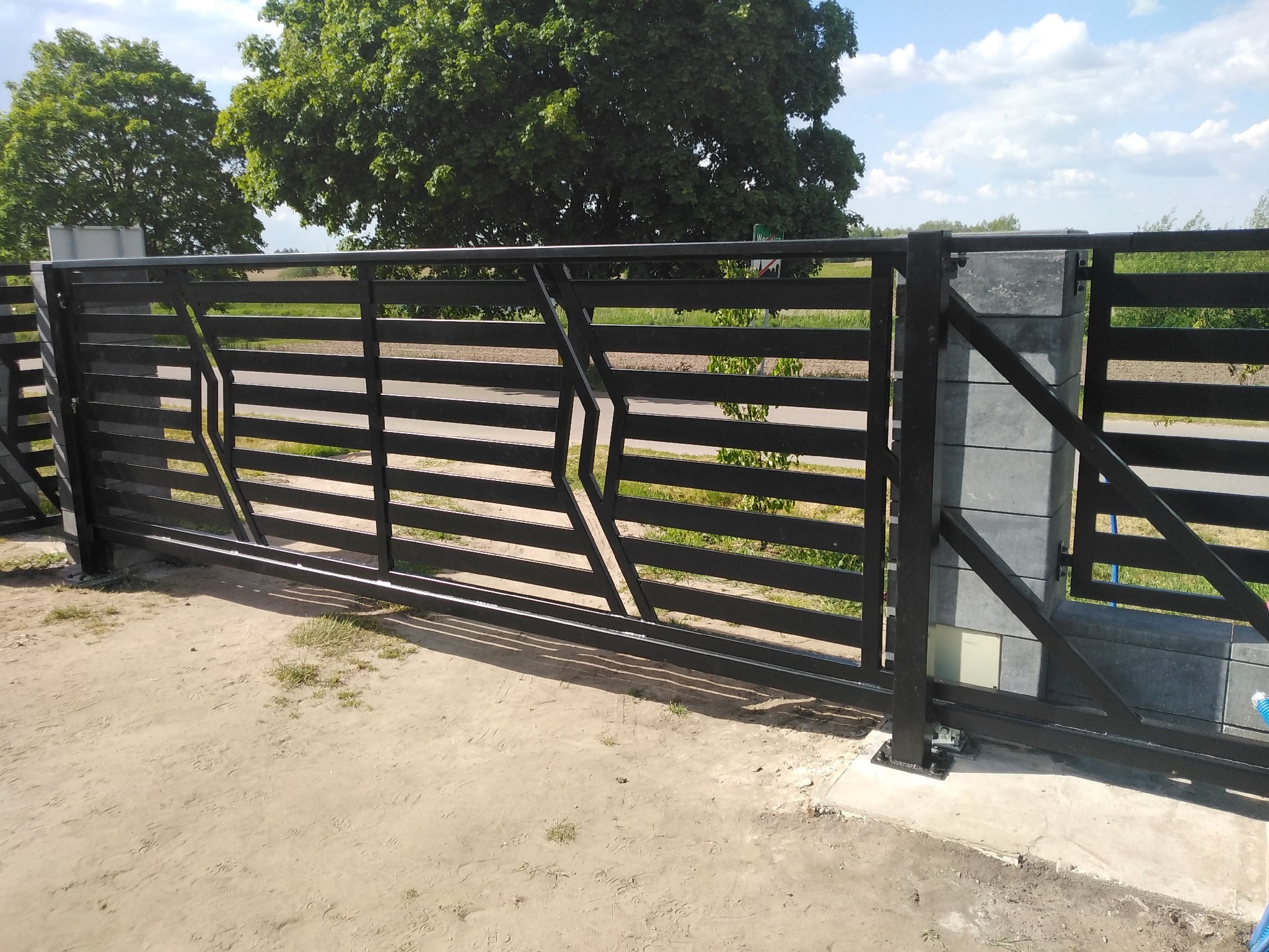 Czarna, przesuwna brama wjazdowa z poziomych profili, zamontowana w Rykach, z widocznym mechanizmem jezdnym i słupkami z szarej kostki.