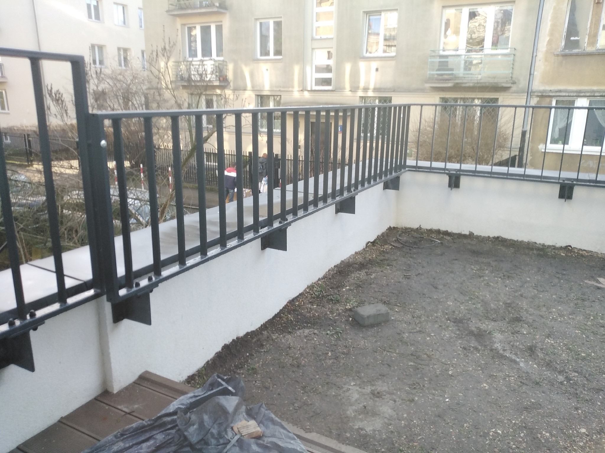 Czarna, metalowa barierka na balkonie z widokiem na budynek mieszkalny, widoczne elementy mocujące do ściany i podłoża, na balkonie ziemia i gruz.