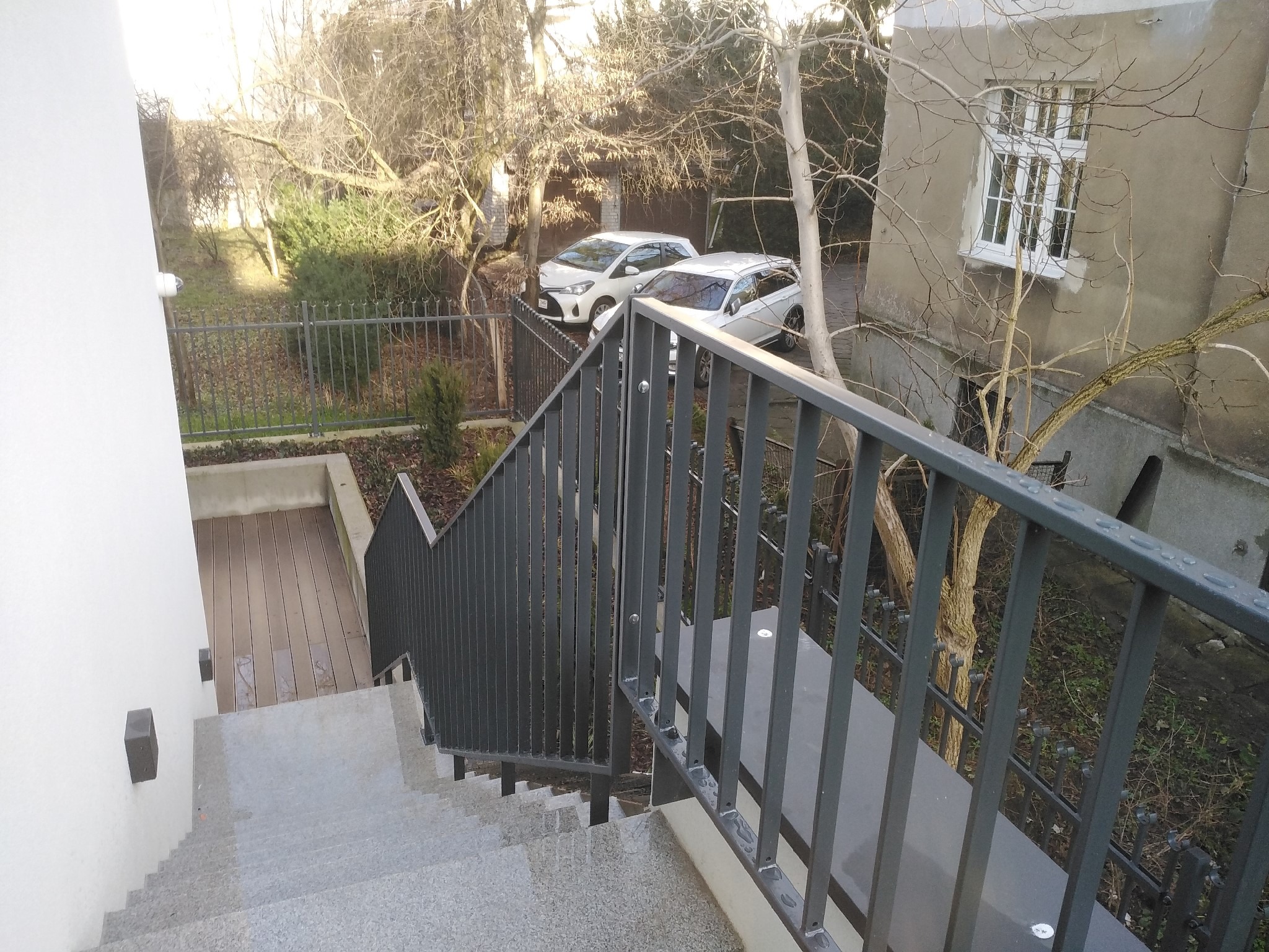 Zewnętrzne schody granitowe z szarą metalową balustradą, widok z góry na dół, w tle dwa samochody i budynek.
