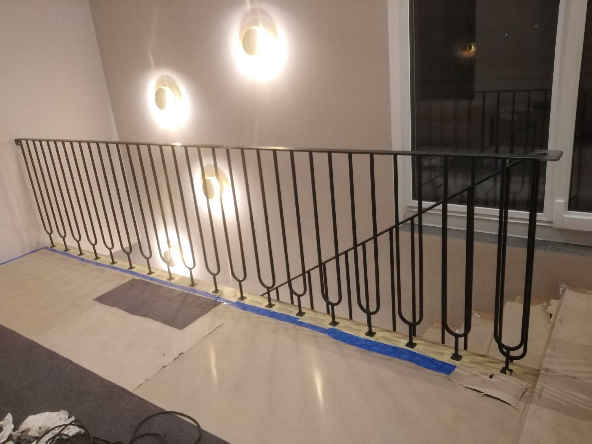Czarna, metalowa balustrada schodowa z charakterystycznymi zaokrąglonymi elementami, zamontowana w jasnym wnętrzu, zabezpieczona niebieską taśmą malarską podczas prac wykończeniowych.