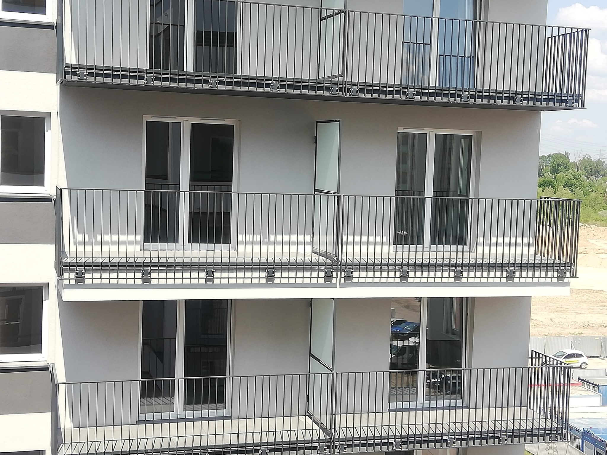 Nowoczesny budynek wielorodzinny z balkonami zabezpieczonymi metalowymi, pionowymi balustradami w kolorze grafitowym. Widoczne otwarte drzwi balkonowe na środkowym piętrze. W tle zieleń drzew i niebo.