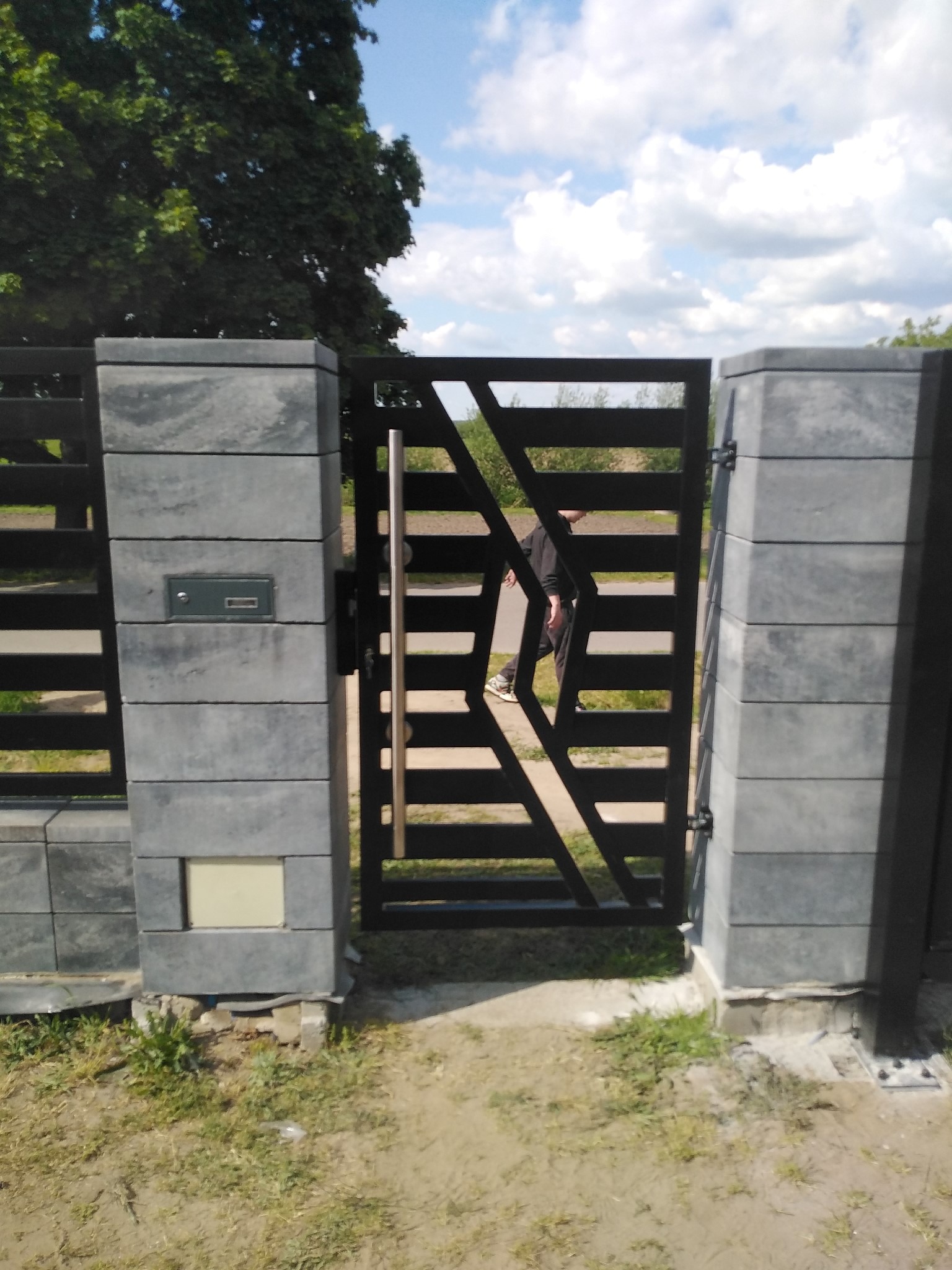 Nowoczesna brama wjazdowa z poziomymi przęsłami i geometrycznym wzorem, osadzona na szarych, betonowych słupkach z zamontowaną skrzynką na listy, widziana z perspektywy podwórza.