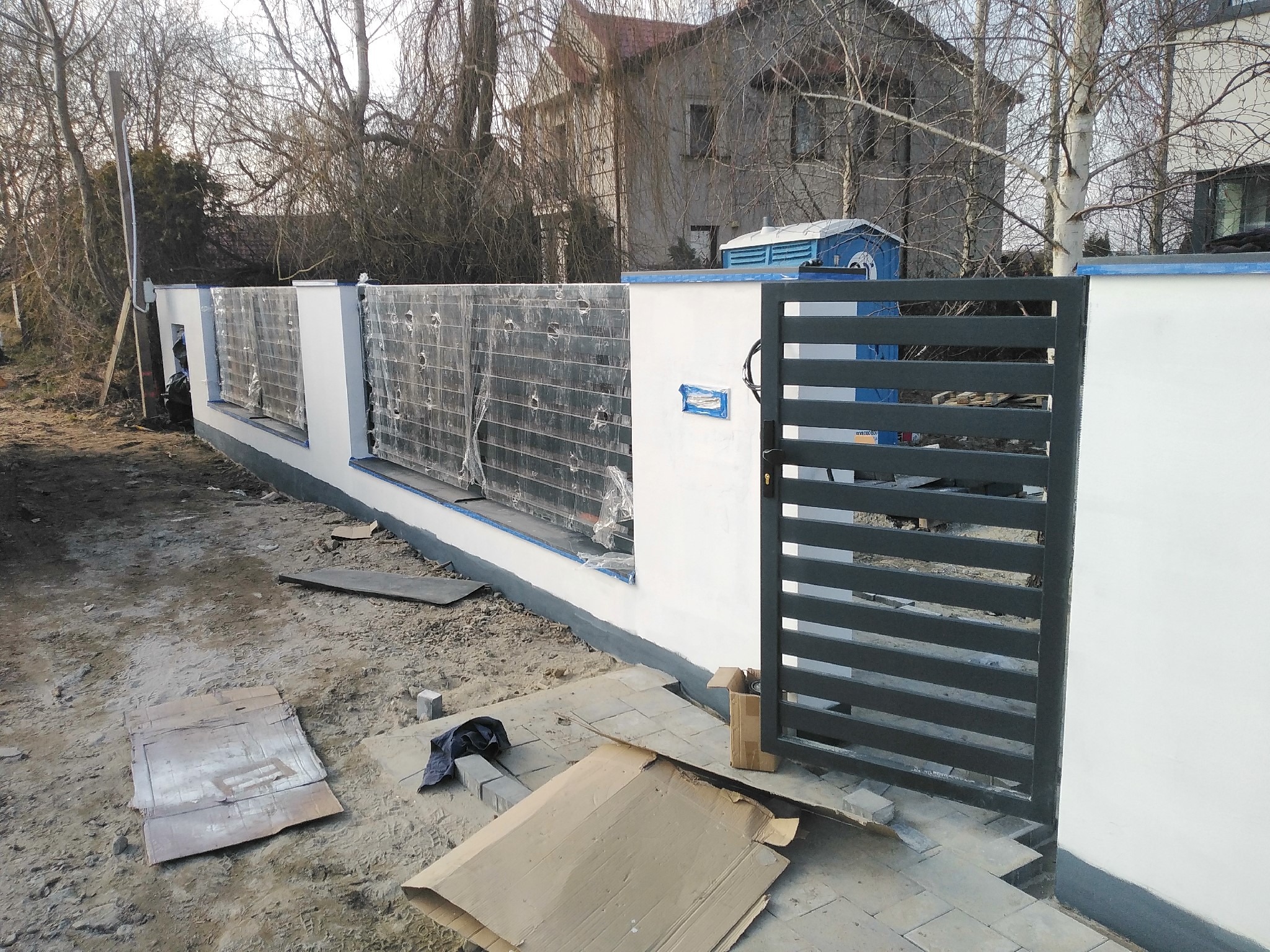Nowoczesne ogrodzenie z bramą w trakcie budowy: białe słupki z szarymi panelami wypełnionymi folią ochronną, szara brama z poziomych profili, widoczne materiały budowlane i teren po budowie.