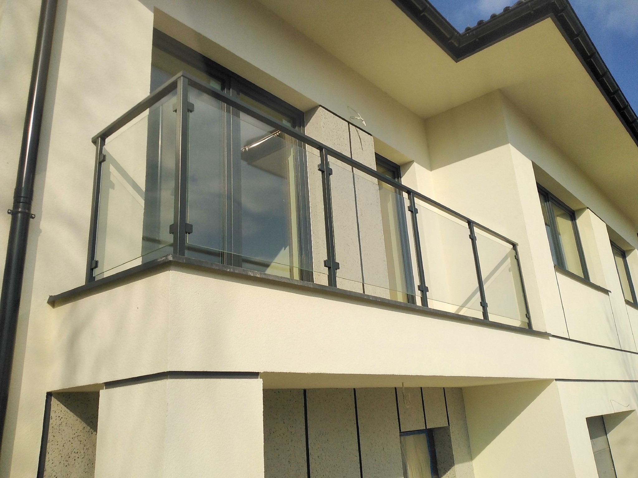 Balkon z nowoczesną balustradą szklaną z czarnymi elementami mocującymi, widoczny z dołu na tle jasnej elewacji budynku.