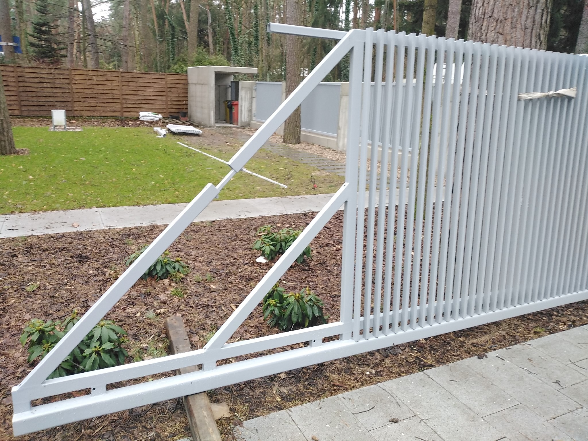 Szara, metalowa brama przesuwna w trakcie montażu, widoczne elementy konstrukcyjne i podkładki stabilizujące, tło z zielenią i elementami architektury ogrodowej.