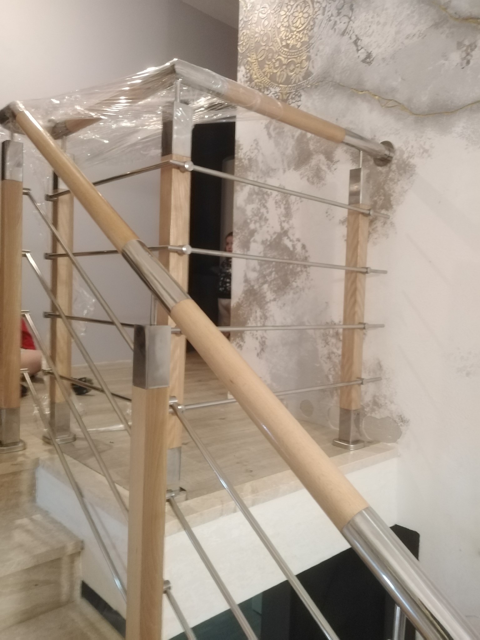 Fragment drewniano-metalowej balustrady schodowej z elementami zabezpieczonymi folią ochronną, widoczne jasne drewniane słupki i stalowe wypełnienie, tło stanowi wzorzysta ściana.