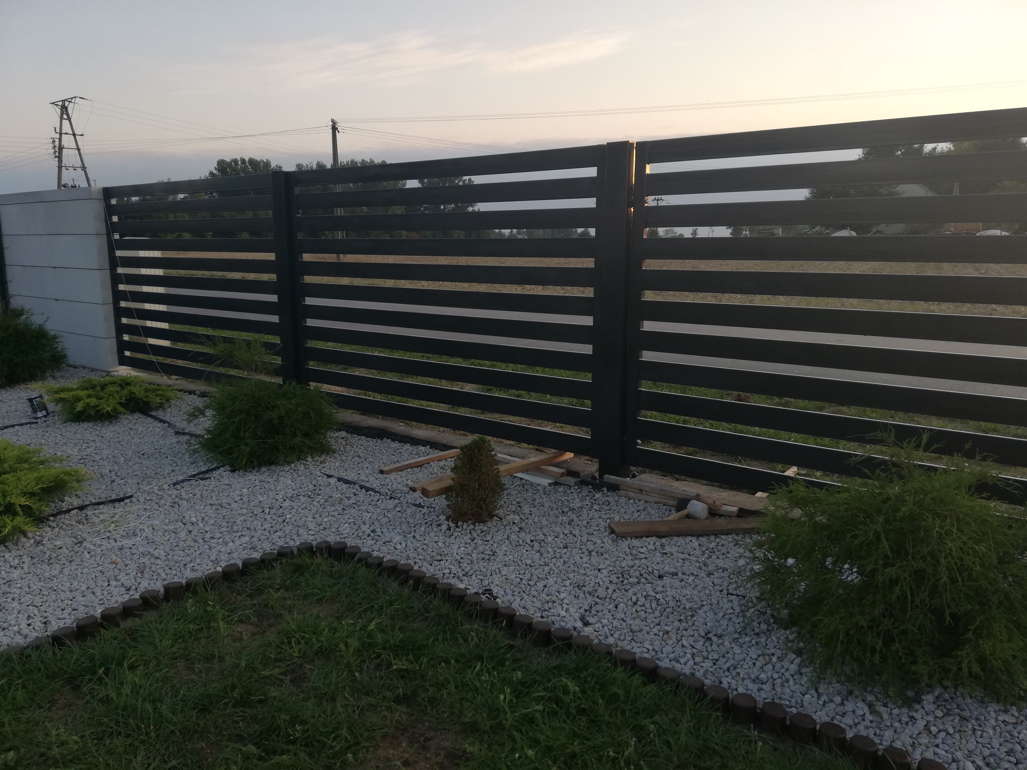 Nowoczesne ogrodzenie panelowe z poziomych, czarnych profili, osadzone na betonowym słupku, otoczone szarym żwirem i niską zielenią, widoczne fragmenty drewnianych elementów montażowych.