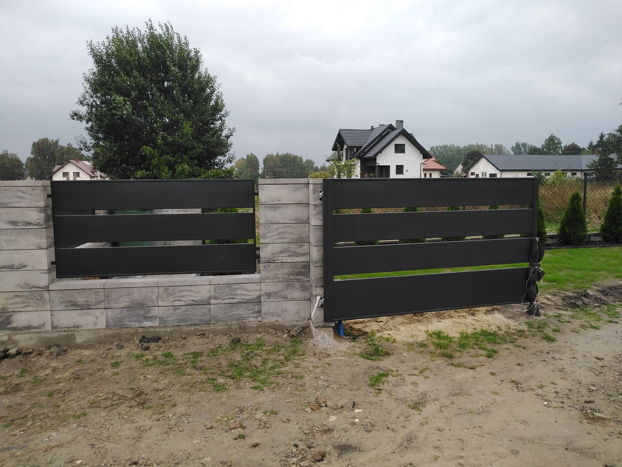 Nowoczesna, pozioma brama wjazdowa i przęsło ogrodzeniowe z szarego metalu, osadzone na betonowych słupkach i podmurówce, na tle domów jednorodzinnych i pochmurnego nieba.