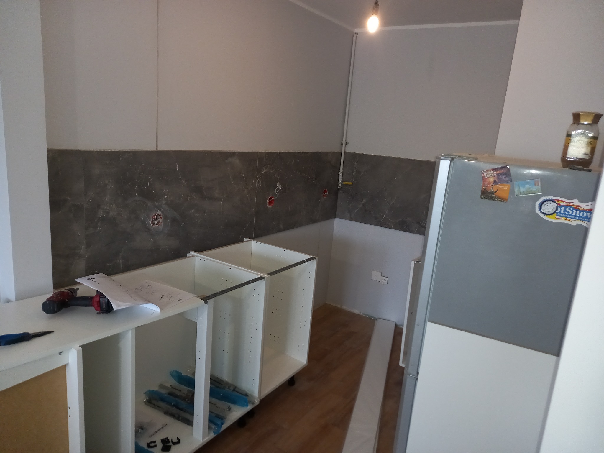 Montaż szafek kuchennych: widoczne niedokończone białe korpusy szafek, w tle szara ściana z marmurowym wzorem, na blacie wiertarka, nóż i instrukcja montażu.