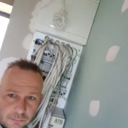 Mężczyzna robiący selfie na tle rozdzielnicy elektrycznej z podłączonymi przewodami w trakcie montażu, widoczne ślady po szpachlowaniu na ścianie.