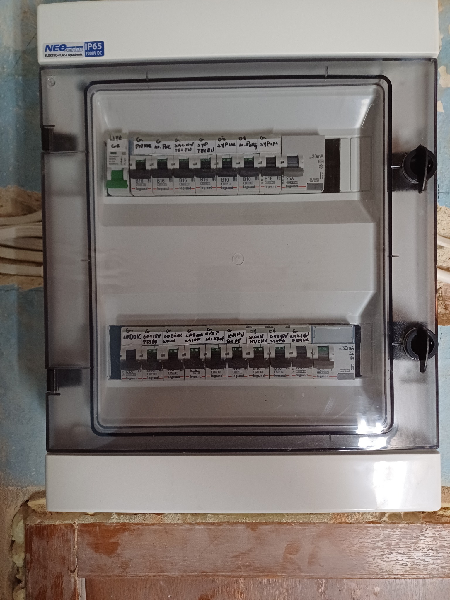 Tablica rozdzielcza z bezpiecznikami, opisana odręcznie, zamontowana w obudowie z tworzywa na ścianie z widocznymi śladami użytkowania.