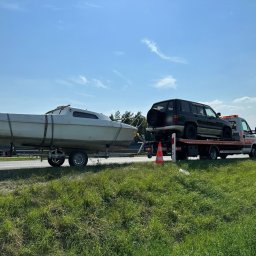 PROMAR Pomoc Drogowa Opole 24h Autostrada A4 Opolskie - Laweta przewozi czarnego SUV-a i białą łódź na przyczepie w słoneczny dzień. Widoczna trawa i niebieskie niebo.